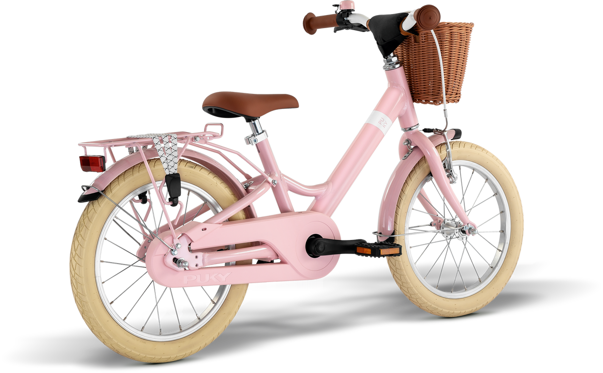 PUKY YOUKE CLASSIC 16 är en rosa barncykel med lättviktsram i aluminium, gräddfärgade däck, brun sadel och styre, flätad korg fram, pakethållare bak och synligt varumärke på ramen. Vit bakgrund.