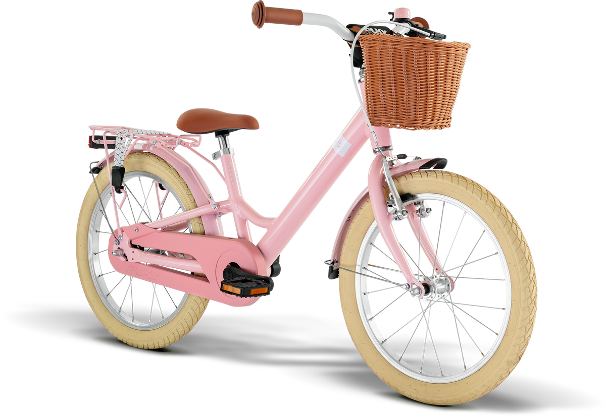 PUKY YOUKE CLASSIC 18 är en rosa cykel med gräddfärgade däck, brun sadel och styre, ett pakethållare bak och en vävd korg fram, mot en vit bakgrund.