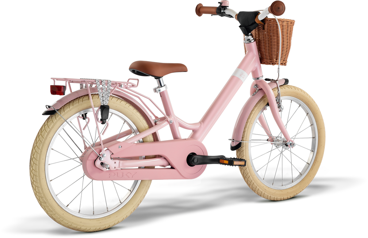 PUKY YOUKE CLASSIC 18 är en rosa barncykel med krämfärgade däck, brun sadel och styre, flätad korg fram och pakethållare med metallskydd bak, mot vit bakgrund.