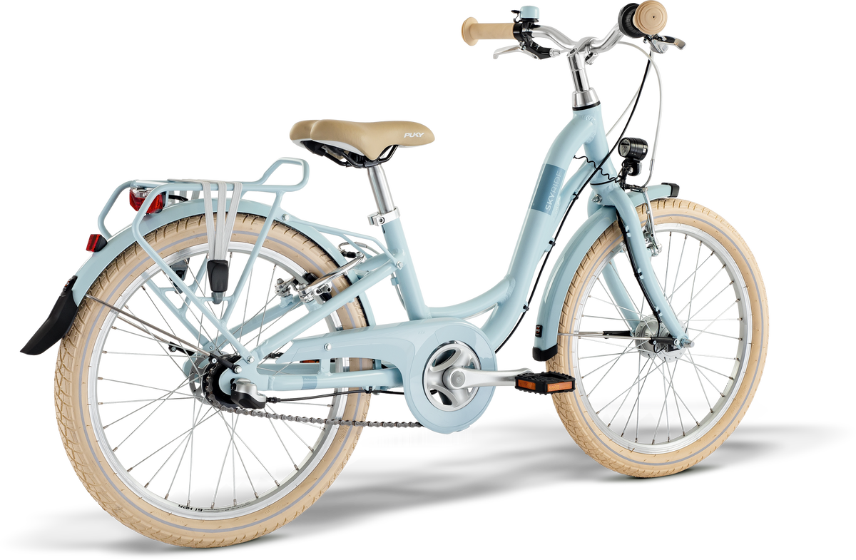 PUKY SKYRIDE 20-3 är en ljusblå cykel med beige däck, solbränd sadel, upprätt styre, pakethållare bak, stänkskärmar, kedjeskydd, fram- och baklyktor. Visas på en vit bakgrund.