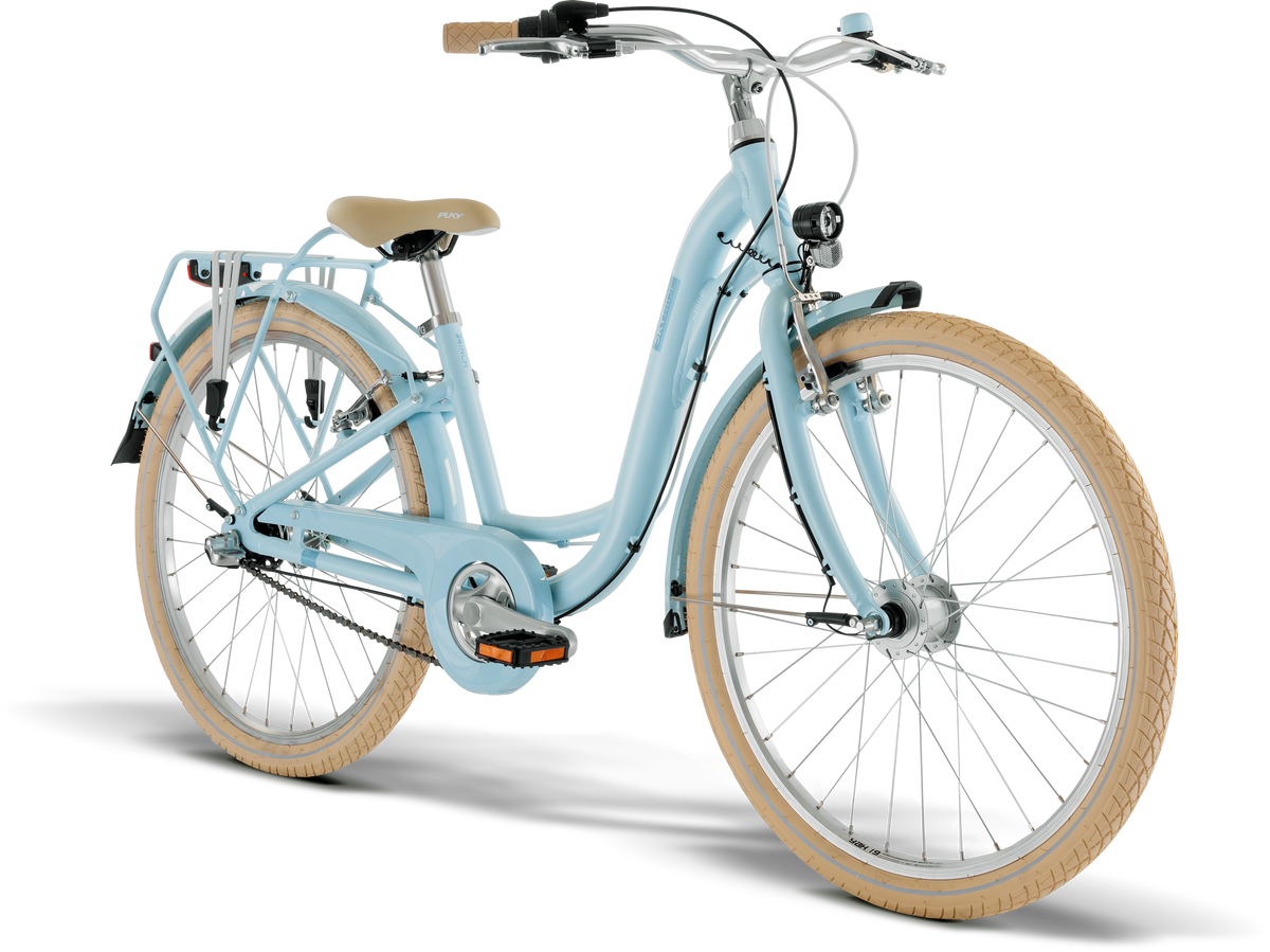 PUKY SKYRIDE 24-3 är en ljusblå genomgångscykel med beige däck och sadel, pakethållare bak, stänkskärmar och strålkastare, mot en vit bakgrund.