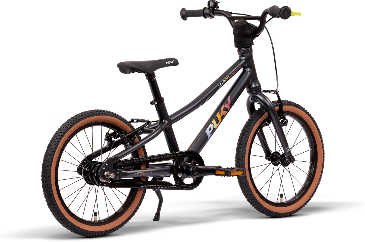 PUKY LS PRO 16-2 FLOWMATIC är en svart barncykel med bruna däck, en svart sadel med märket "PUKY", ett rakt styre med gult ändlock på höger handtag och ett stödben, som visas mot en vit bakgrund.