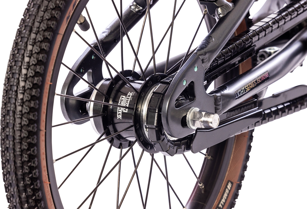 Närbild av PUKY LS PRO 16-2 FLOWMATIC-cykelns bakhjul med däckmönster, svarta ekrar, nav, kedja och rambultar och beslag mot en vit bakgrund.