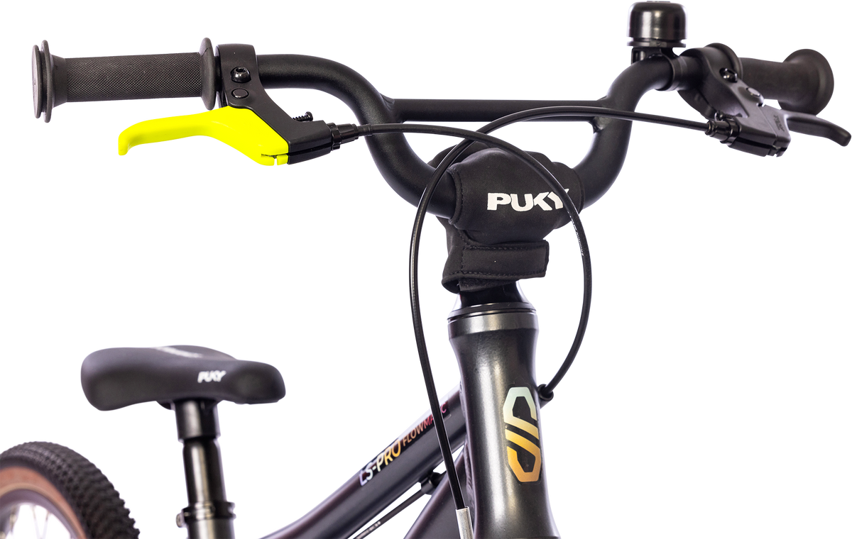 Närbild av ett svart PUKY LS PRO 16-2 FLOWMATIC styre med en gul bromshandtag till vänster, en ringklocka till höger och ett vadderat headsetskydd. Överdelen av sadeln och framramen på denna PUKY-cykel är också synliga.
